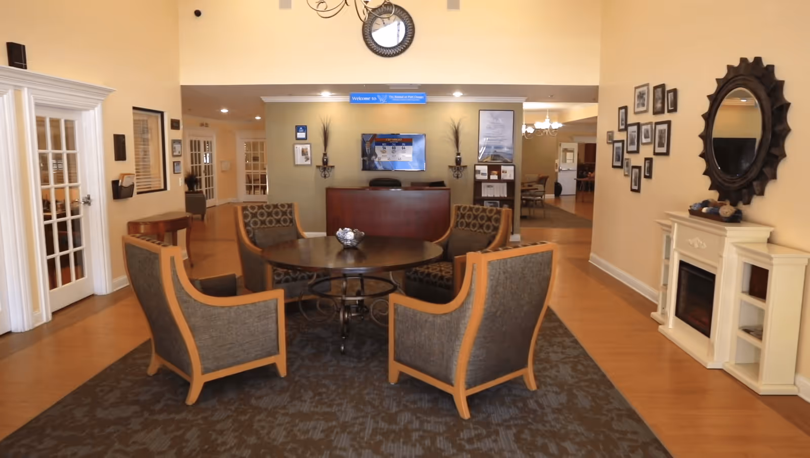 Interior view of a senior living facility common area with four cushioned armchairs arranged around a round wooden table on a patterned carpet. In the background, there is a reception desk with a TV mounted on the wall above it displaying a weather forecast. The walls are decorated with framed pictures, a large round mirror, and a white electric fireplace. The space has warm lighting and wooden flooring.