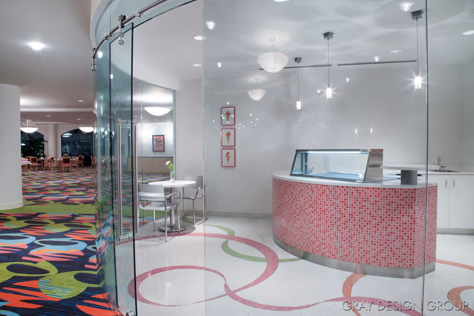 Interior dining area with a curved glass entrance, a pink-tiled curved service counter, tables, and colorful patterned carpeting.