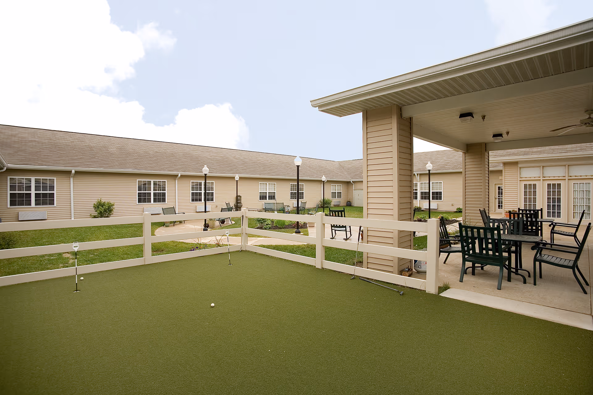 Outdoor courtyard area at Waterford Crossing featuring a small fenced putting green, patio with tables and chairs, surrounded by single-story beige buildings with multiple windows under a partly cloudy sky.
