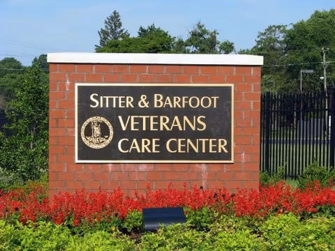 A brick sign with a black plaque that reads 'Sitter & Barfoot Veterans Care Center' surrounded by red flowers and green shrubs, with trees and a black metal fence in the background under a clear blue sky.