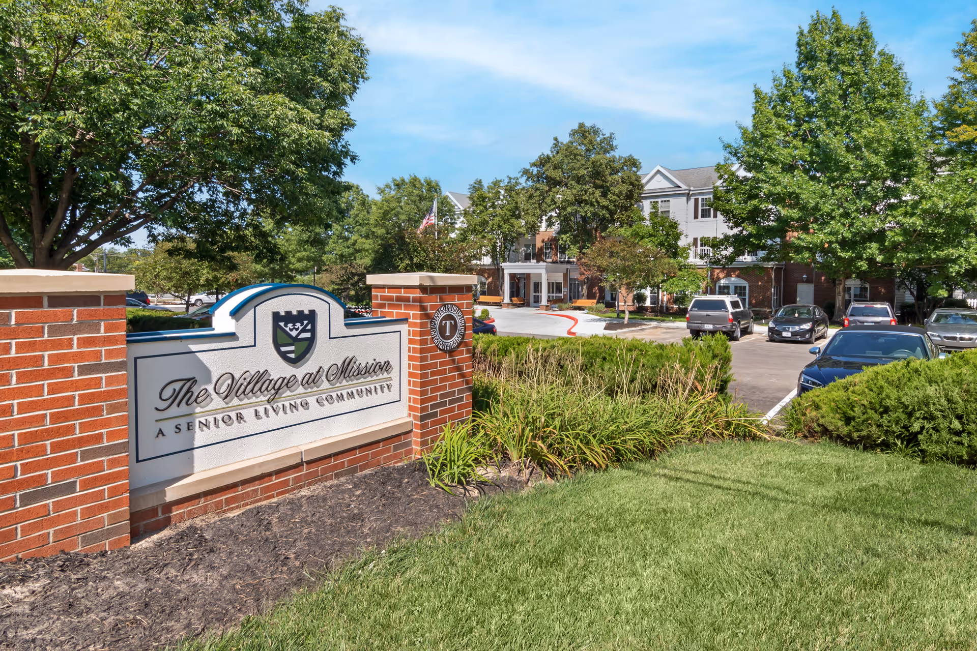 Entrance sign for The Village at Mission, a senior living community, with a brick and stone structure surrounded by green grass, bushes, and trees. In the background, there is a parking lot with several cars and a multi-story building under a blue sky.