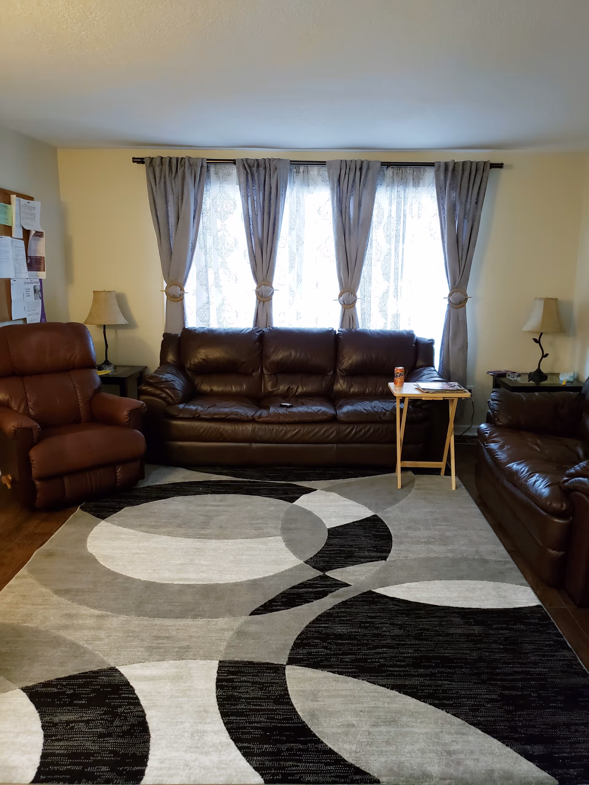 A living room with three brown leather seating pieces including a sofa, loveseat, and recliner. There is a patterned area rug with black, gray, and white circular designs on the floor. Behind the sofa, there is a window with sheer white curtains and gray drapes tied back. Two side tables with lamps are positioned on either side of the seating area. A small wooden tray table holds a can of soda and some magazines.