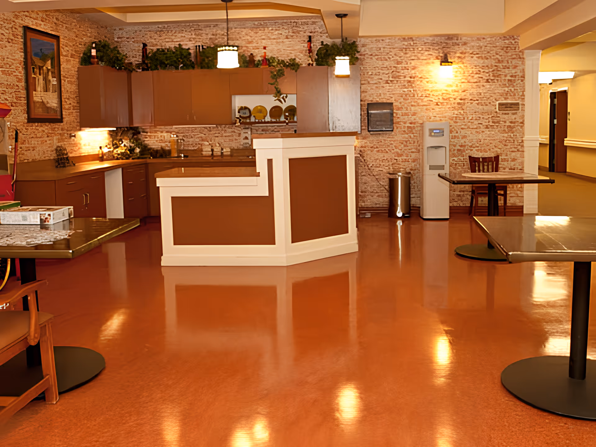 Interior view of a communal area in a senior living facility featuring a reception or service counter with brown and white paneling, brown cabinets along a brick wall, hanging pendant lights, a water dispenser, and several tables and chairs on a polished brown floor.