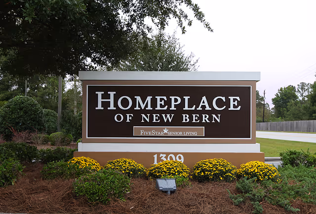 Entrance sign reading "Homeplace of New Bern" surrounded by landscaping and yellow mums.