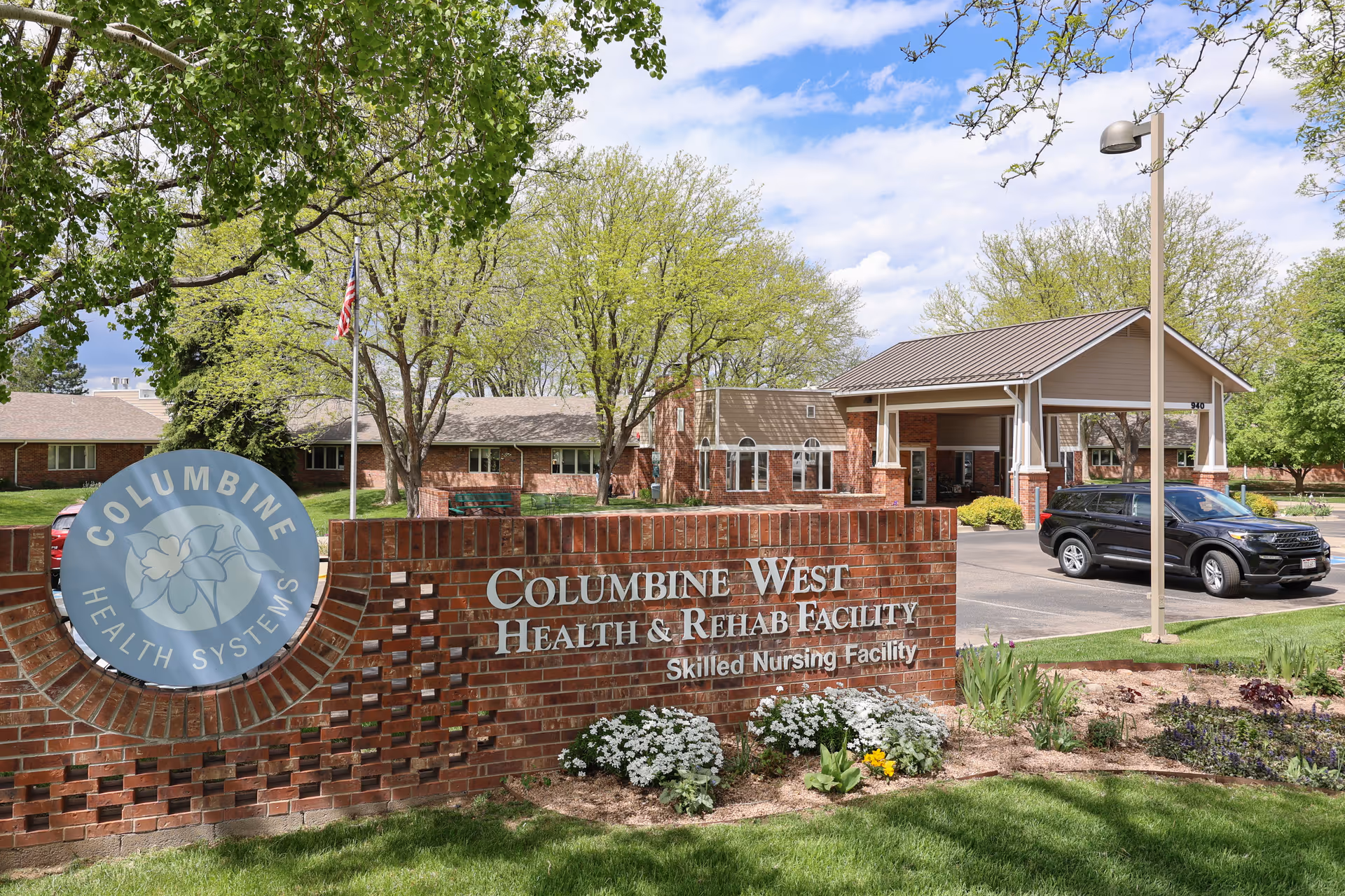 Exterior view of Columbine West Health & Rehab Facility showing a brick sign with the facility's name and logo, a driveway with a black SUV parked, and trees with green leaves under a partly cloudy sky.