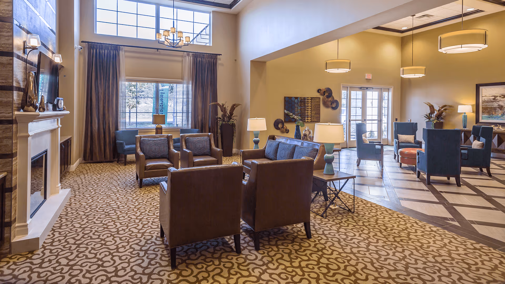 A spacious and well-lit living room area in a senior living facility featuring multiple seating arrangements including leather armchairs and blue upholstered chairs. The room has large windows with curtains, a fireplace with decorative items on the mantel, patterned carpet, and modern hanging light fixtures. There are also side tables with lamps and decorative wall art.