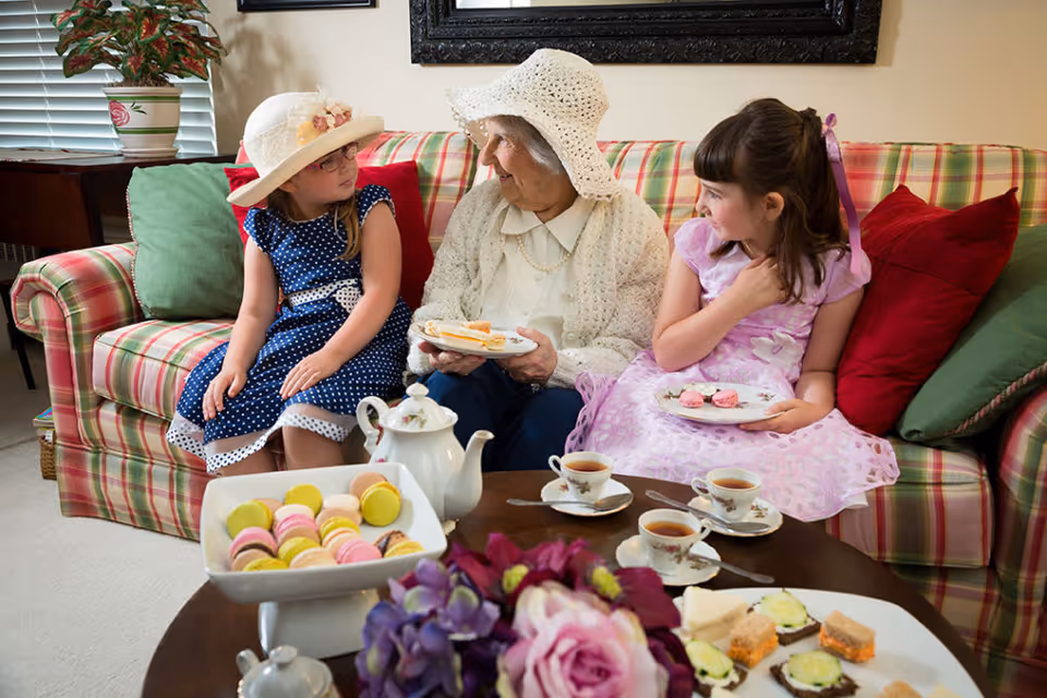 An elderly woman wearing a white crocheted hat and sweater sits on a plaid couch between two young girls dressed in fancy dresses and hats. They are having tea with plates of sandwiches, macarons, and tea cups on a wooden table in front of them. The setting is a cozy living room with colorful cushions and a potted plant near a window with blinds.