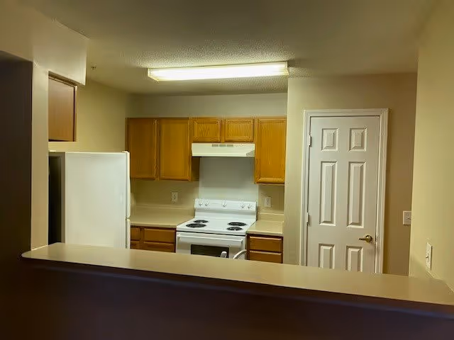 Interior view of a kitchen with wooden cabinets, a white refrigerator, a white electric stove with an overhead range hood, beige countertops, and a closed white door to the right.