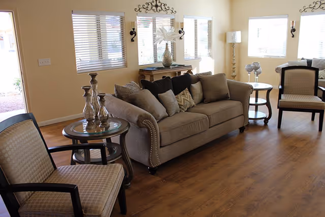 A bright living room with wooden flooring featuring a beige sofa adorned with multiple cushions, two patterned armchairs, and two round glass-top side tables with decorative items. The room has several windows with blinds allowing natural light to fill the space, and a decorative wall piece above a console table with a vase and other decor.