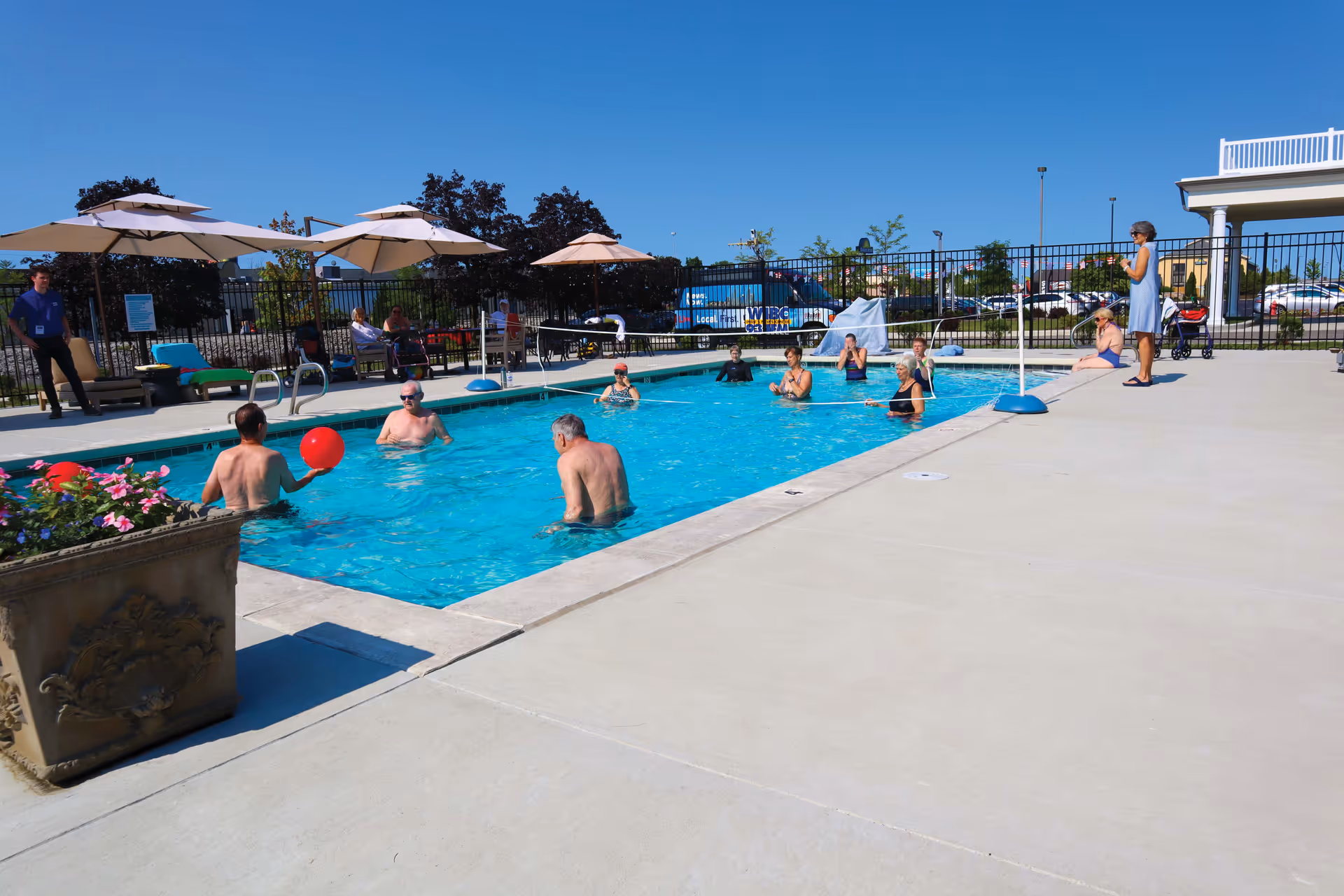 Several elderly people enjoying a sunny day in an outdoor swimming pool, some standing in the water and others sitting by the poolside. There are umbrellas and lounge chairs around the pool, with a clear blue sky overhead and a fence enclosing the area.