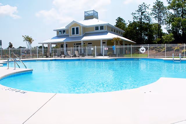 Outdoor swimming pool with clear blue water in front of a two-story building with a white roof and a wraparound porch, surrounded by a white fence and some trees in the background under a partly cloudy sky.