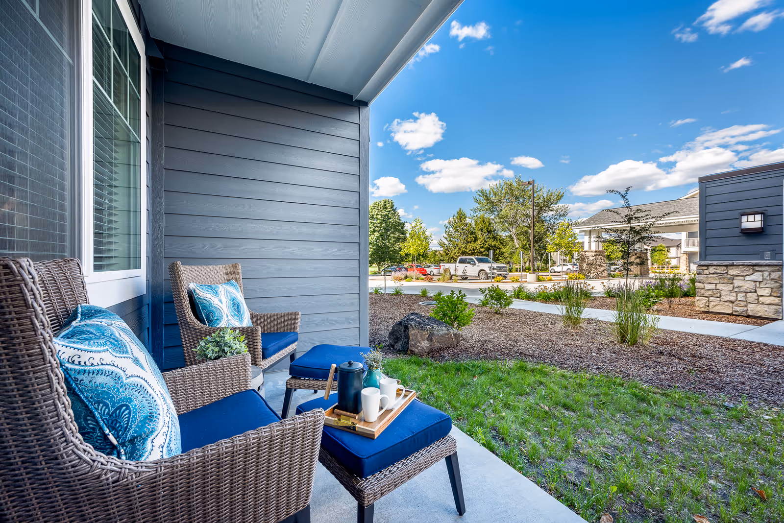 A cozy outdoor patio area with two wicker chairs and a matching ottoman, all with blue cushions and decorative pillows. A tray with a coffee pot and cups is placed on the ottoman. The patio overlooks a landscaped garden area with grass, shrubs, and a rock, with a parking lot and building structures visible in the background under a blue sky with scattered clouds.