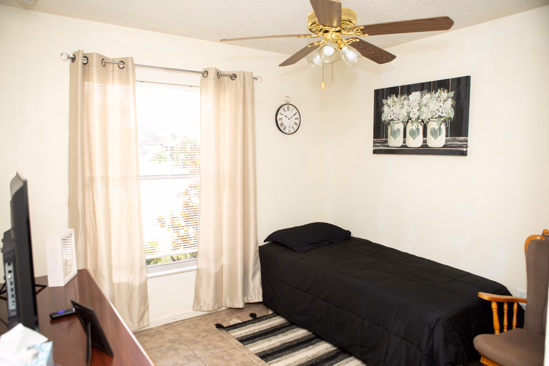 A small bedroom with a single bed covered in a black comforter and pillow. There is a window with beige curtains, a ceiling fan with lights, a wall clock, and a painting of flowers in jars on the wall. A wooden chair with a cushion and a dresser with a TV and some items are also visible.