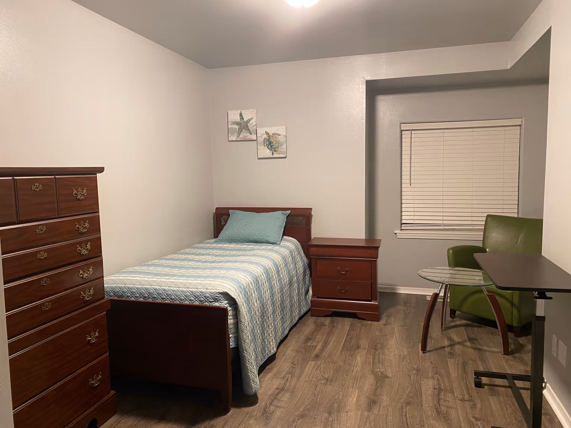A small bedroom with a single bed covered in a blue and white striped quilt and a matching pillow. Next to the bed is a wooden nightstand with two drawers. On the left side of the room is a tall wooden dresser with multiple drawers. A green armchair and a glass-top side table are positioned near a window with closed blinds. The floor is wood, and the walls are painted light gray with two small sea-themed paintings hanging above the bed.