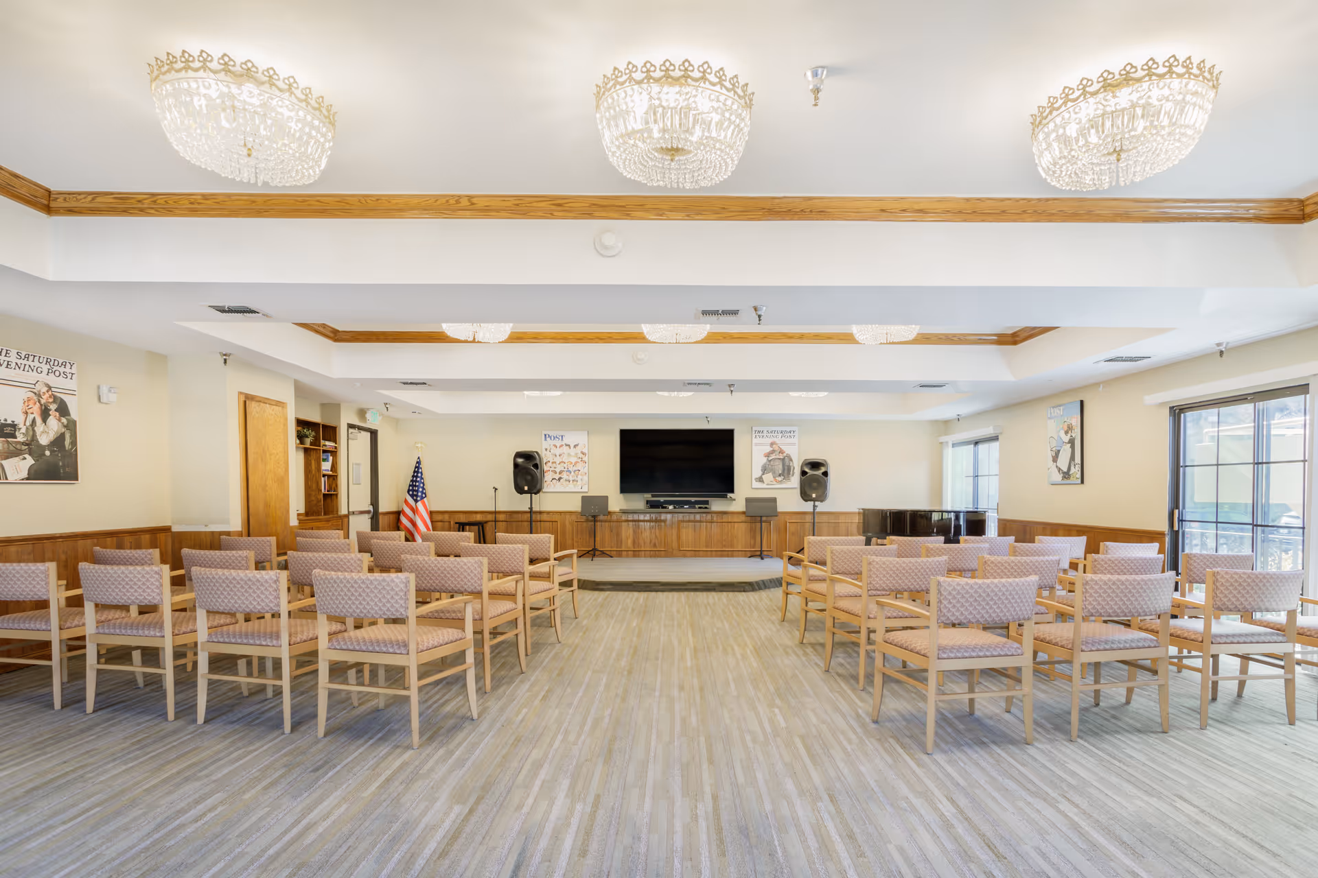 A bright multipurpose meeting room with rows of wooden chairs facing a small stage, TV, speakers, and an American flag.