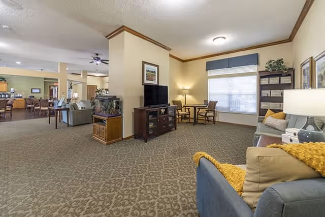 A spacious and well-lit common area in a senior living facility featuring a patterned carpet, a TV on a wooden stand, a fish tank, a small table with two chairs near a window with blinds, and several seating options including sofas with cushions and throws. The room has soft yellow walls and ceiling lights, with some framed artwork and plants for decoration.