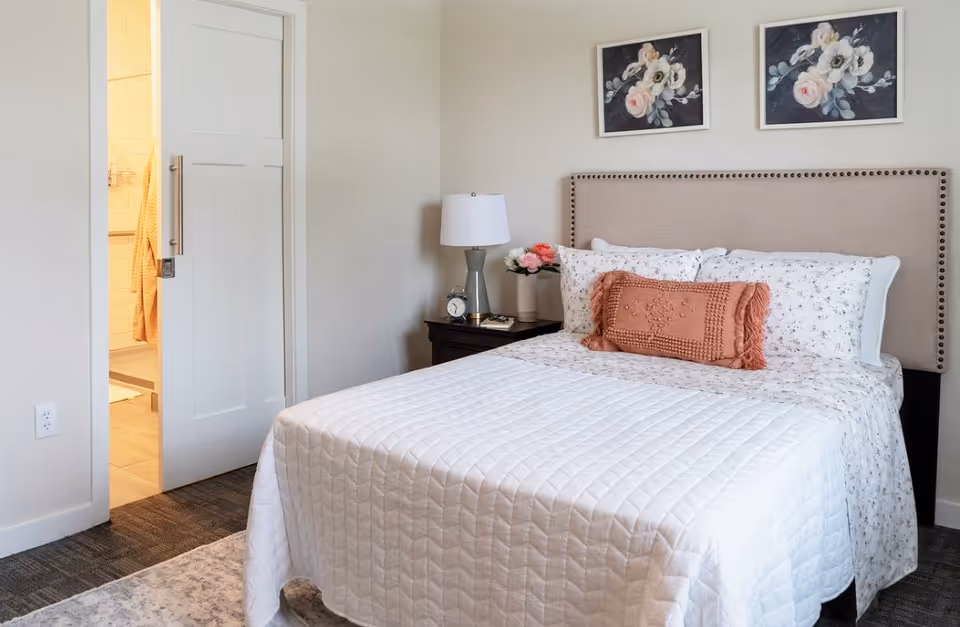 A neatly made bed with a beige upholstered headboard, white quilted bedspread, floral sheets, and a decorative rust-colored pillow. On the right side of the bed is a dark wooden nightstand with a white lamp, a clock, and a vase of flowers. Two floral paintings hang on the wall above the bed. To the left, a partially open white door reveals a glimpse of a bathroom with beige tiles and a hanging towel.