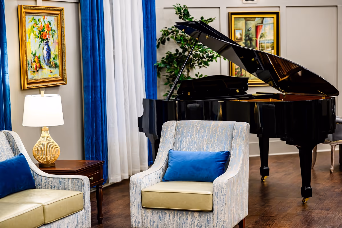 A cozy living room area featuring a black grand piano, two upholstered armchairs with blue cushions, a wooden side table with a lamp, blue and white curtains, and framed paintings on the walls.