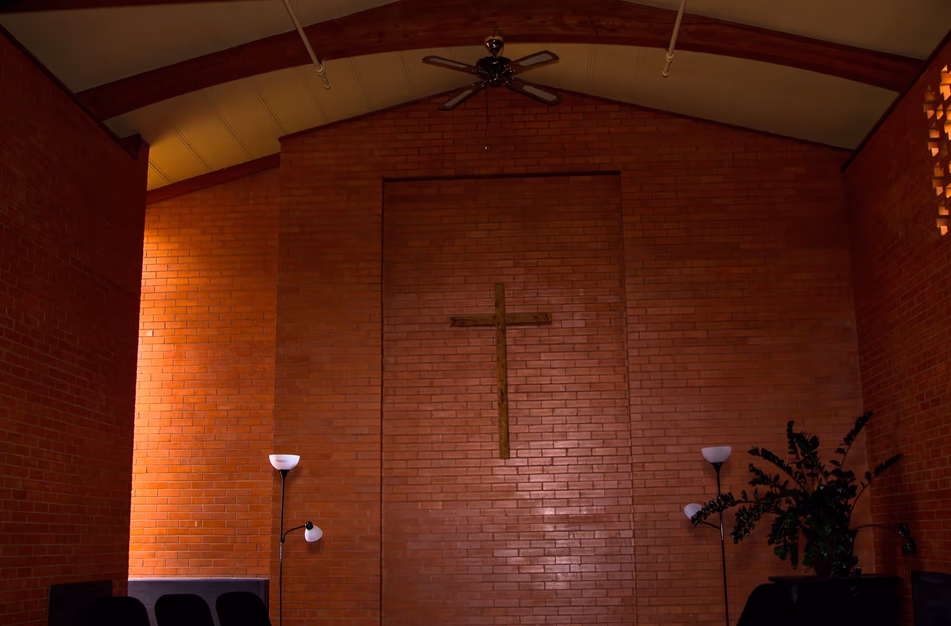 Interior view of a room with brick walls and a wooden cross mounted on the central wall. The ceiling is vaulted with a ceiling fan. There are floor lamps and some plants visible at the sides of the room.
