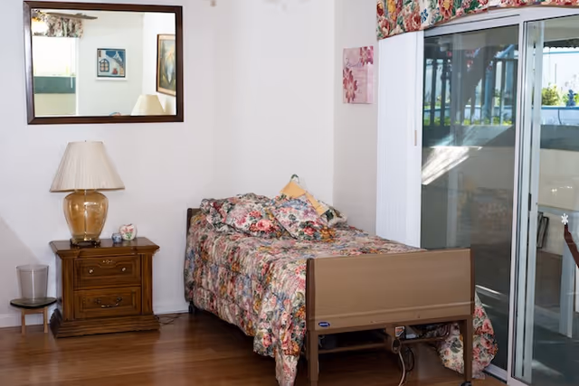 A small bedroom with a single bed covered in a floral bedspread and matching pillows. Next to the bed is a wooden nightstand with a lamp and small decorative items. A large mirror hangs on the wall above the nightstand. To the right, there is a sliding glass door leading to an outdoor area with a patio table and chairs visible outside.