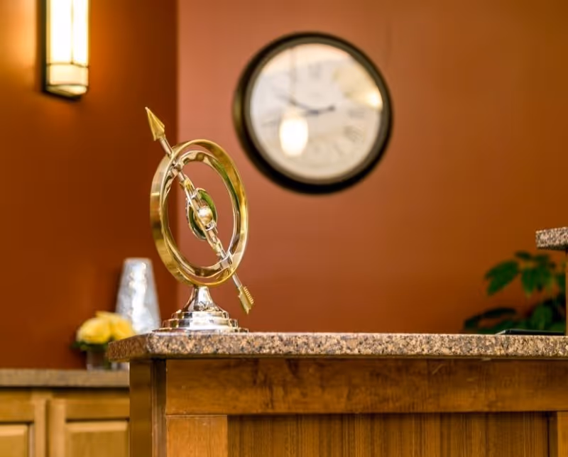 A decorative metallic armillary sphere with an arrow sits on a granite countertop in a room with warm brown walls. A round wall clock with Roman numerals and a wall sconce light are visible in the background, along with a small plant and a silver vase with yellow flowers.