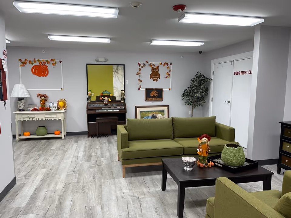 A cozy common area in Sandalwood Village featuring green sofas and a black coffee table with decorative items. The room has light gray walls and wood-patterned flooring. On the back wall, there is a piano with a mirror above it, autumn-themed decorations including a pumpkin and an owl, and a small table with a lamp and more fall decorations. A potted plant is placed near a white door with a sign that reads 'DOOR MUST CL'. The ceiling has bright fluorescent lights.