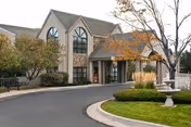 Exterior view of a senior living facility building with large arched windows, a stone and stucco facade, surrounded by landscaped greenery including trees and bushes, and a curved driveway leading to the entrance.