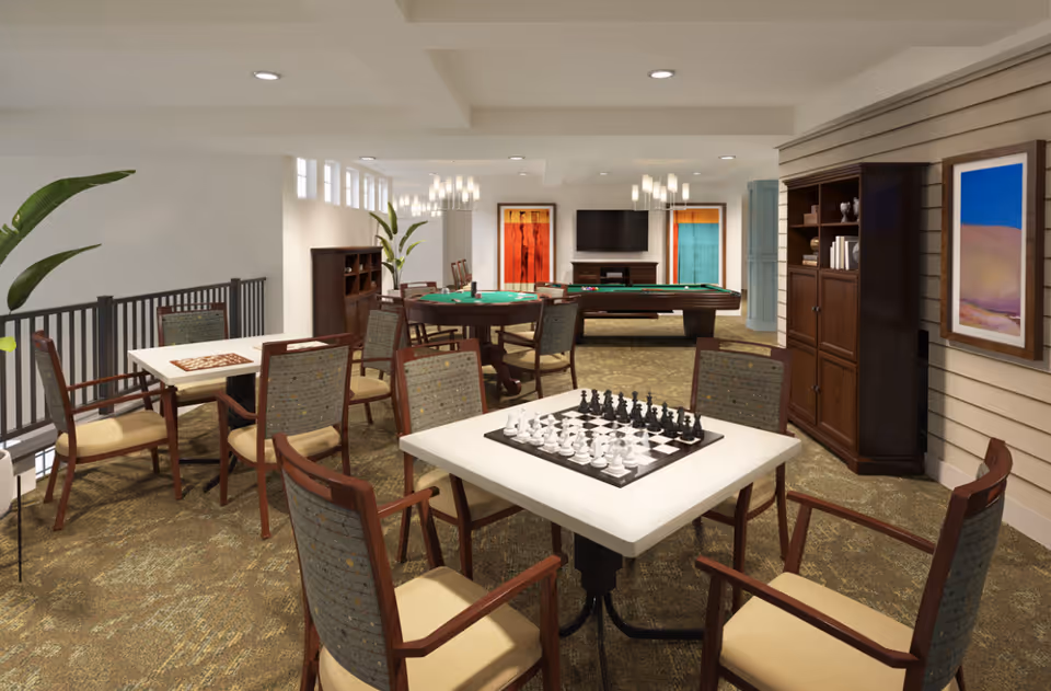 A well-lit game room with multiple tables and chairs. One table has a chessboard set up, another has a card game setup, and a third table has a green felt top for card games. In the background, there is a pool table and a flat-screen TV mounted on the wall. The room features carpeted flooring, framed artwork, plants, and wooden cabinets for storage.