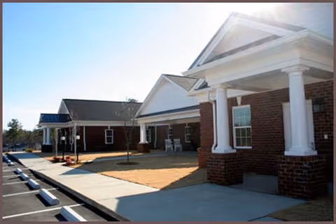 Exterior view of a single-story brick building with white columns and a covered porch area, adjacent to a parking lot with marked spaces under a clear blue sky.