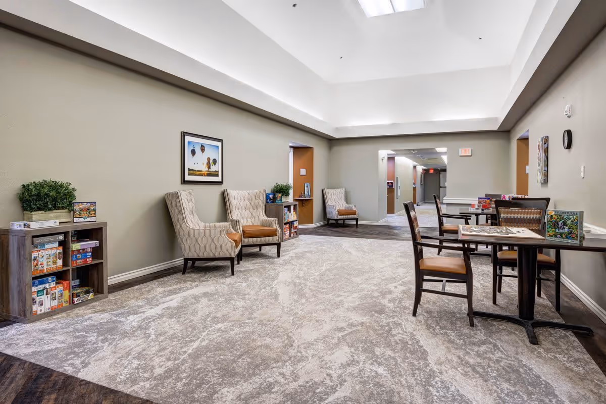 A spacious, well-lit common area with a high ceiling and skylights. The room features comfortable armchairs along the wall, small tables with chairs, and shelves filled with board games and puzzles. The walls are painted light gray, and there is a framed picture of hot air balloons hanging on one wall.