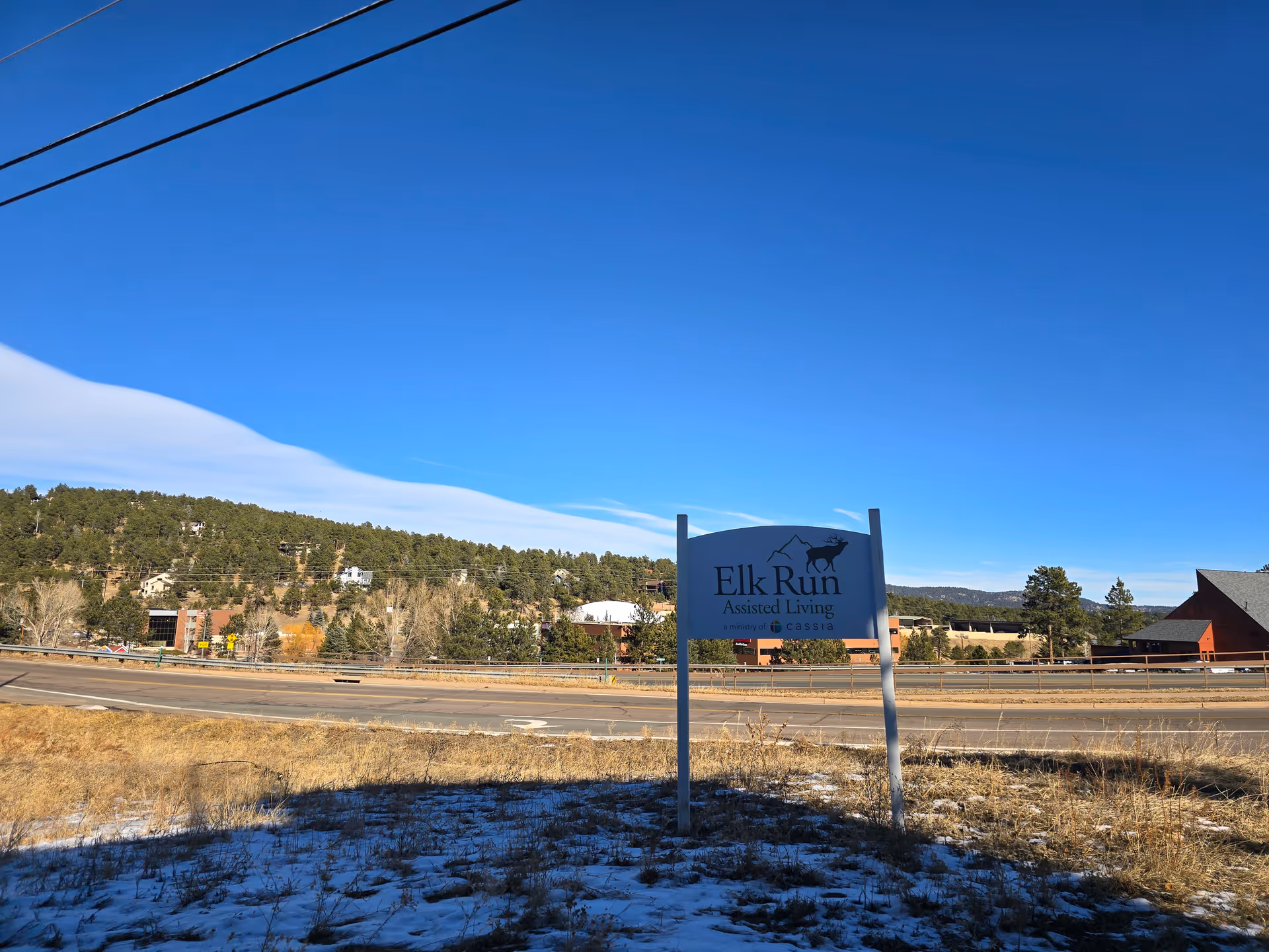 Outdoor scene showing a sign for Elk Run Assisted Living with a clear blue sky, some snow on the ground, a road, trees, and buildings in the background.