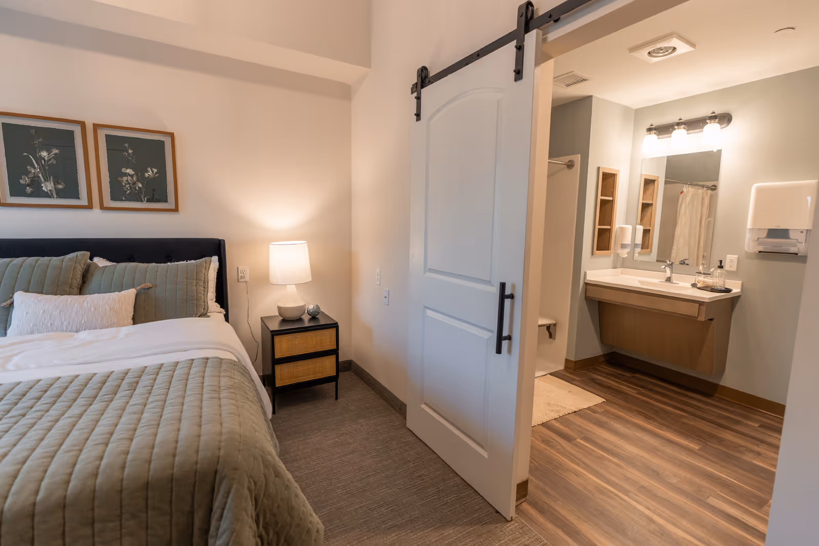 A cozy bedroom with a neatly made bed featuring green and white pillows and a matching quilt. A bedside table with a lamp is next to the bed. A sliding barn door is partially open, revealing a bathroom with a sink, mirror, light fixture, and a shower area with a curtain.