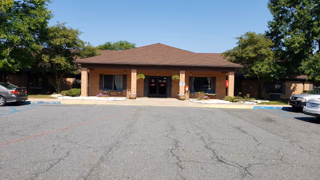 Front exterior view of a single-story brick building with a brown shingled roof, two large windows with curtains on either side of a double glass door entrance, surrounded by small plants and trees, with a parking lot in front under a clear blue sky.