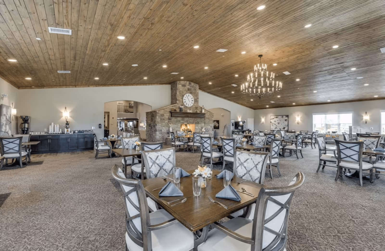 Spacious dining room with multiple tables and chairs arranged neatly. Each table is set with glasses, napkins, and silverware. The room features a high wooden ceiling with recessed lighting and a large chandelier. A stone fireplace with a clock above it is centrally located, and large windows allow natural light to fill the space.