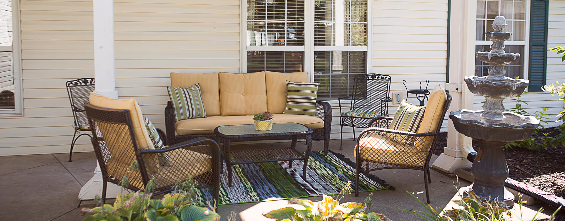Outdoor patio area with cushioned wicker chairs and a sofa arranged around a glass-top coffee table on a striped outdoor rug, next to a multi-tiered stone water fountain and a building with beige siding and windows.