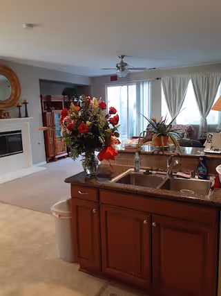 Open-plan kitchen island with a sink and a large vase of flowers overlooking a living room with a fireplace and sliding glass doors.