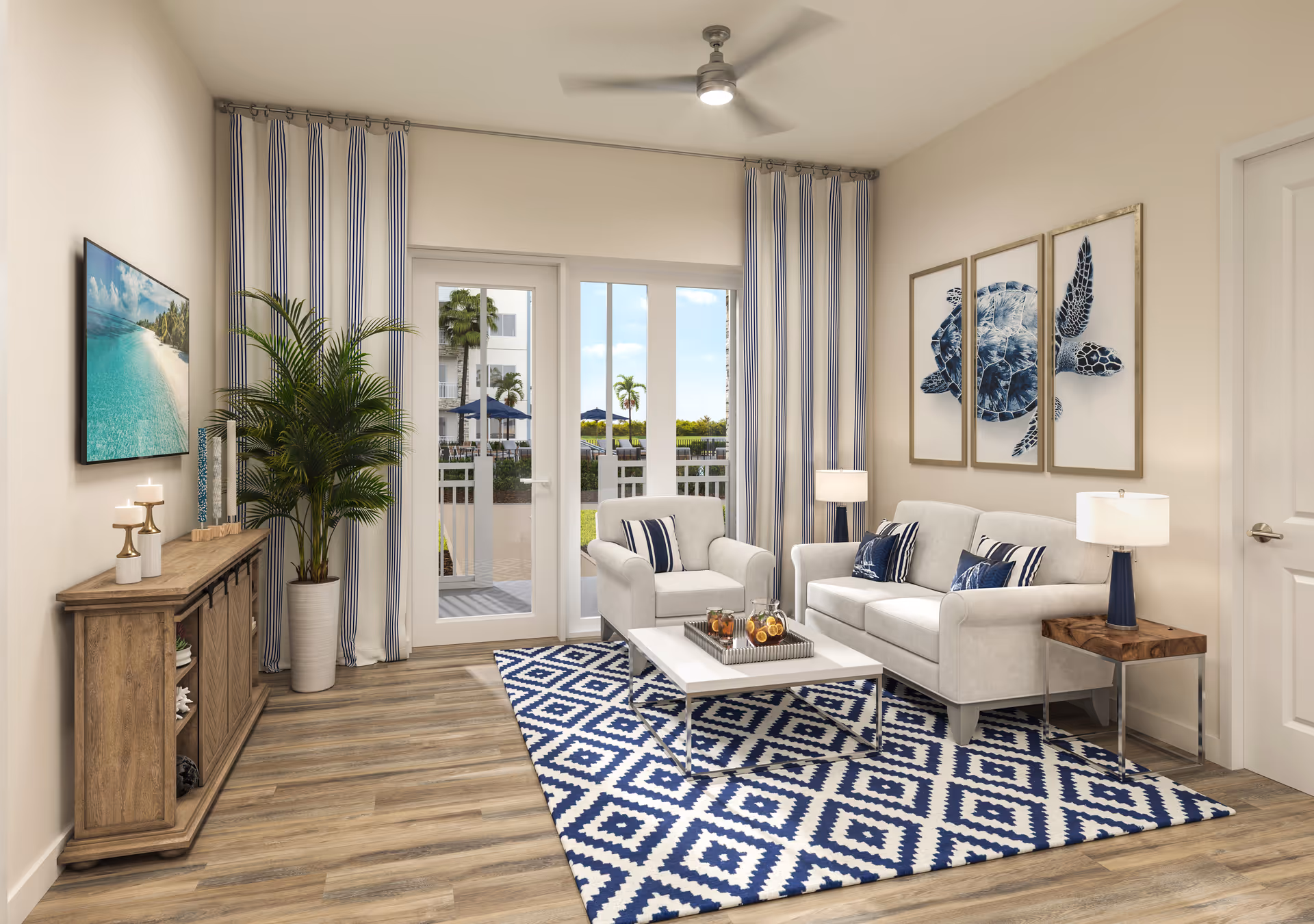 A bright and modern living room with light-colored walls and wood flooring. The room features a white sofa and armchair with blue and white patterned cushions, a white coffee table on a blue and white geometric rug, and two side tables with lamps. There is a large potted plant next to a wooden console table with candles and decorative items. Blue and white striped curtains frame glass doors that open to an outdoor area with palm trees. Three framed pictures of a sea turtle hang on the wall above the sofa.