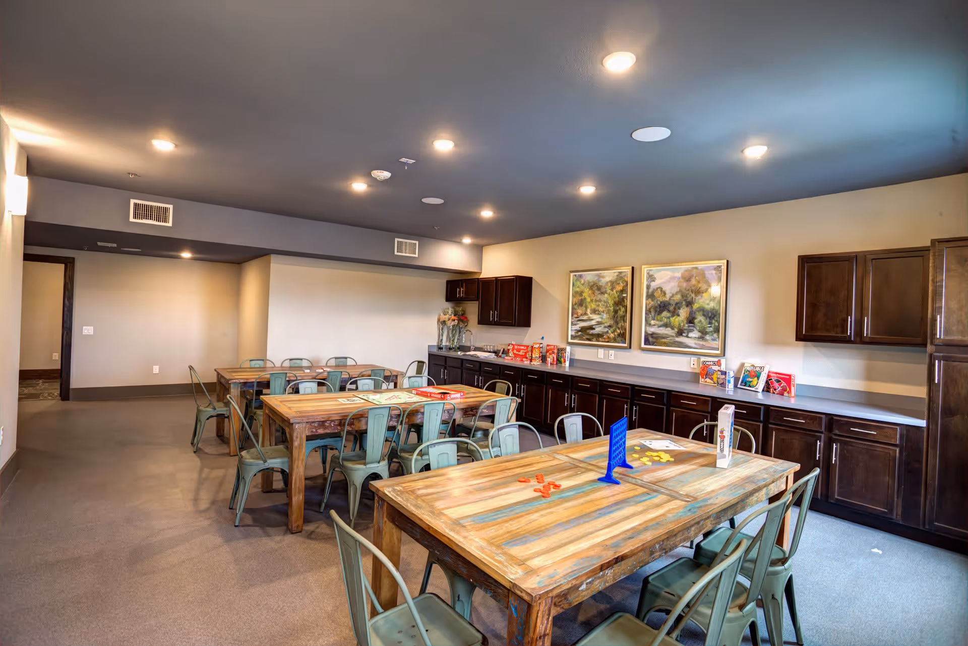 A well-lit activity room with wooden tables and metal chairs. The tables have board games and game pieces on them. The room has beige walls, a dark ceiling with recessed lighting, and dark wooden cabinets along one wall. Two framed landscape paintings hang above the cabinets.