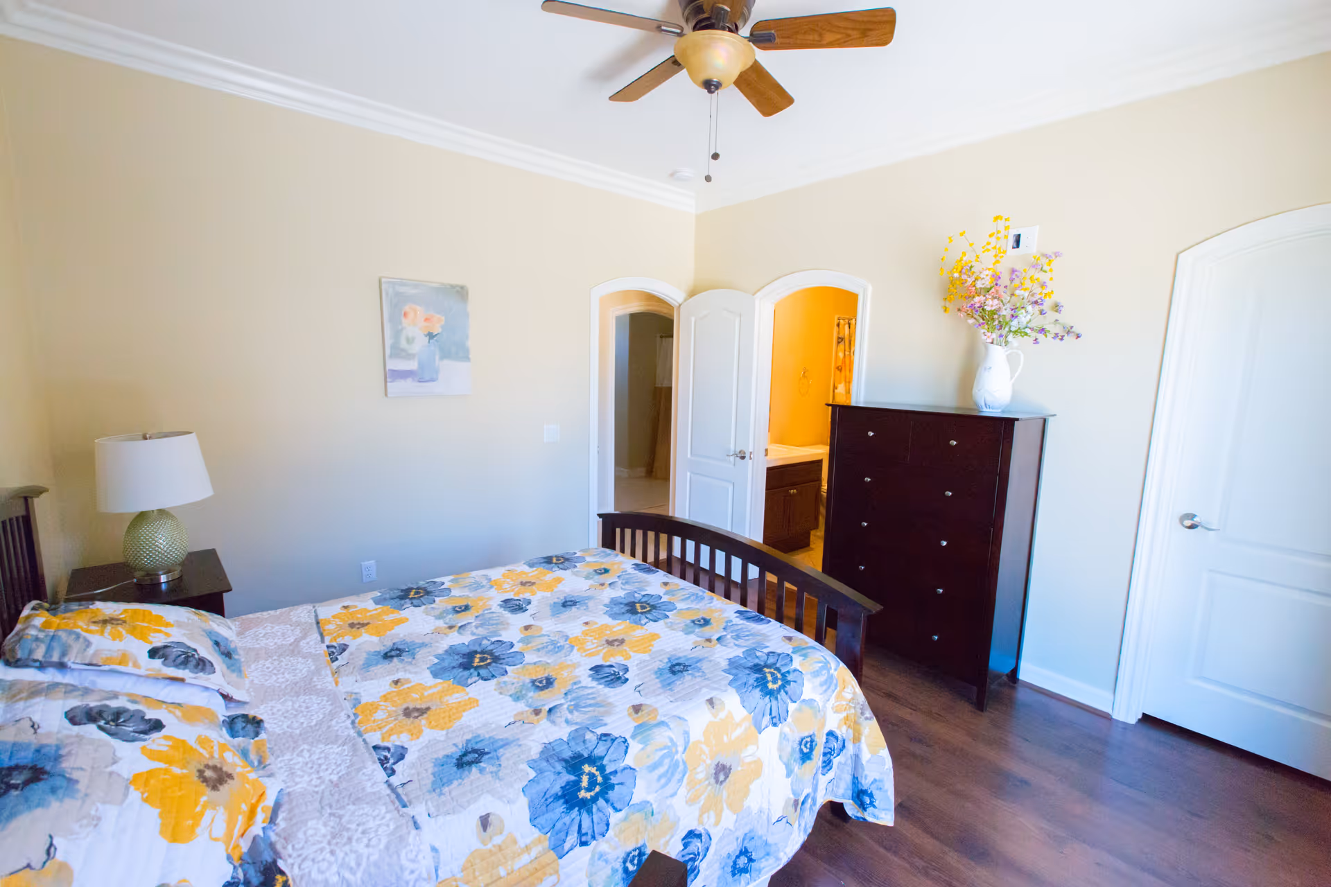 A bright bedroom with a wooden bed featuring a floral bedspread in yellow, blue, and gray colors. There is a nightstand with a lamp on the left side of the bed, a dark wooden dresser with a vase of flowers on top, and two white doors, one leading to a bathroom with a visible sink and shower curtain. The room has light beige walls, a ceiling fan with wooden blades, and wooden flooring.