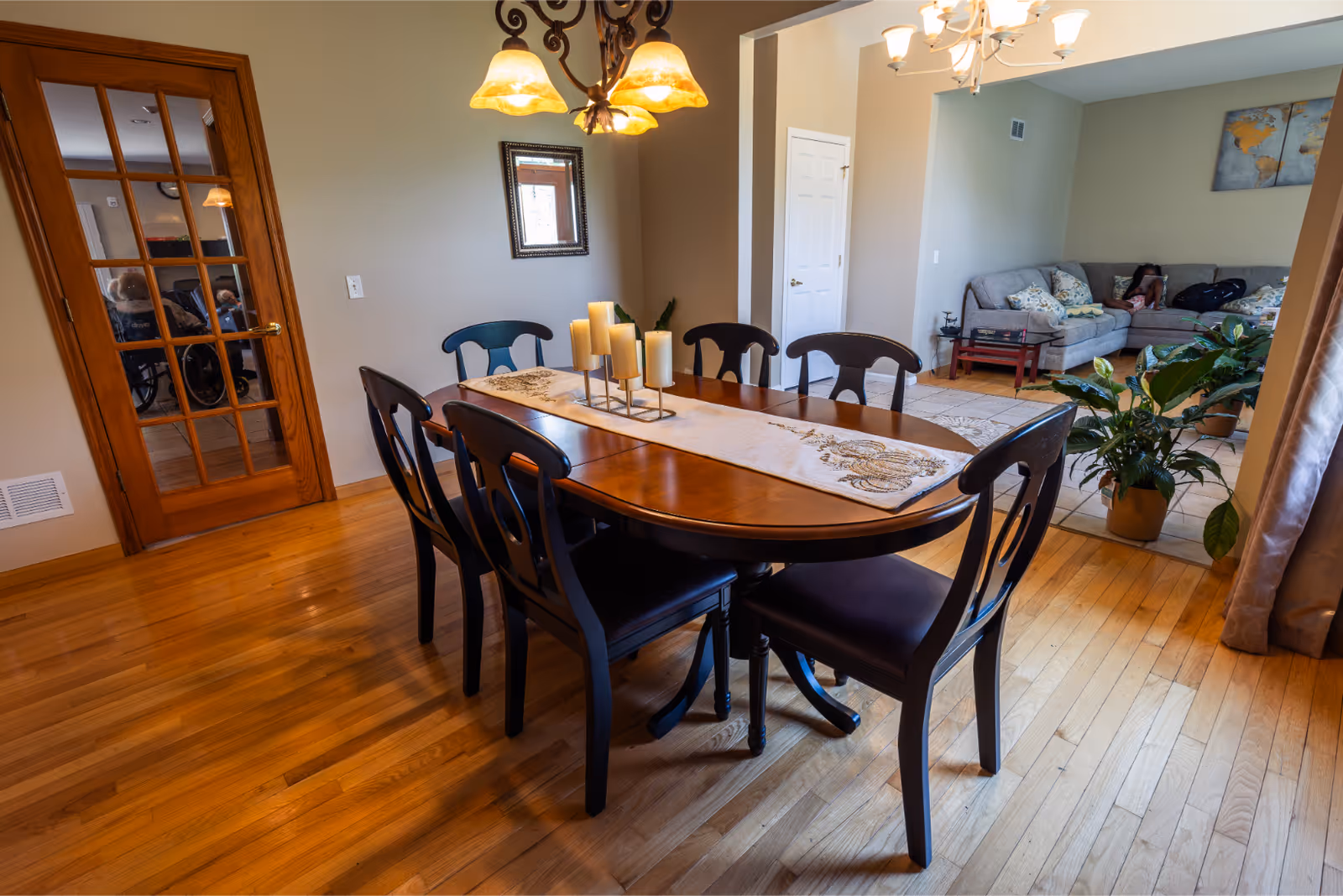 A dining room with a wooden table and six black chairs. The table has a decorative runner and a candle centerpiece. In the background, there is a living room area with a gray sectional sofa, a small table, and a large plant. The floor is wooden, and there is a glass-paneled wooden door on the left side.