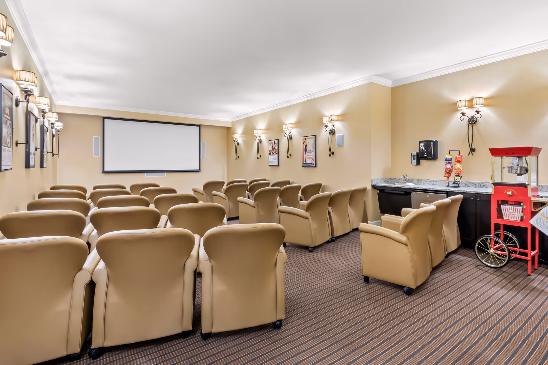 A cozy movie theater room with multiple rows of beige leather armchairs facing a large blank projection screen. The walls are painted light beige and decorated with framed movie posters and wall sconces. On the right side, there is a countertop with a popcorn machine and a snack rack.