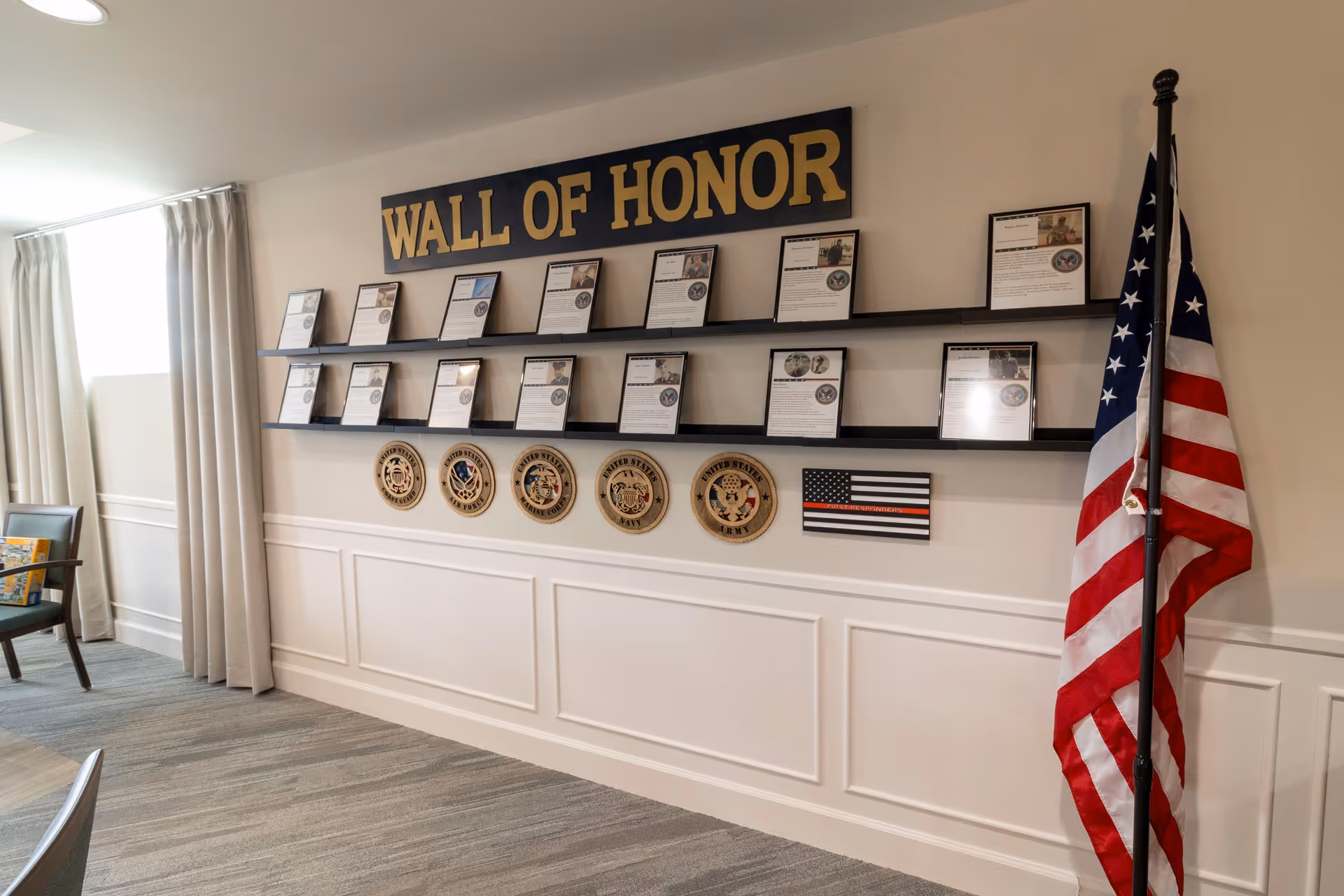 Interior view of a wall display titled 'Wall of Honor' featuring framed photos and descriptions of individuals, military branch emblems, and an American flag on a stand to the right. The room has light-colored walls, carpeted flooring, and a window with curtains on the left side.