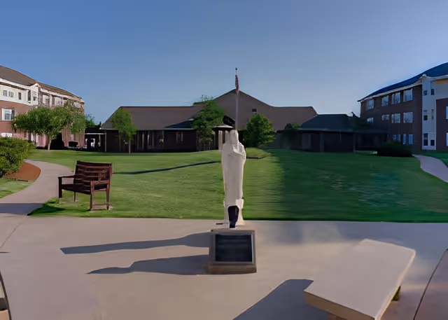 Outdoor courtyard area at Saint Ann Retirement Center featuring a paved walkway, a wooden bench, a modern abstract sculpture on a pedestal, green lawns, and buildings in the background under a clear blue sky.