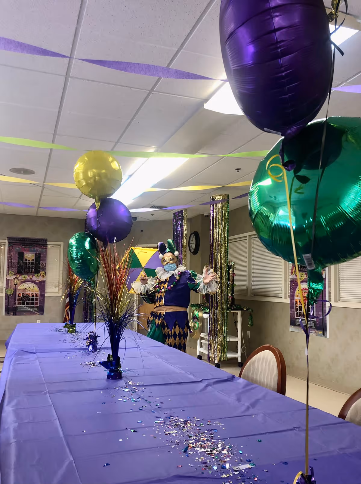 A decorated room with a long table covered in a purple tablecloth, adorned with colorful confetti and centerpieces with balloons in purple, green, and yellow. Streamers hang from the ceiling. In the background, a person dressed in a colorful jester costume and mask stands near the wall, which has Mardi Gras-themed decorations.