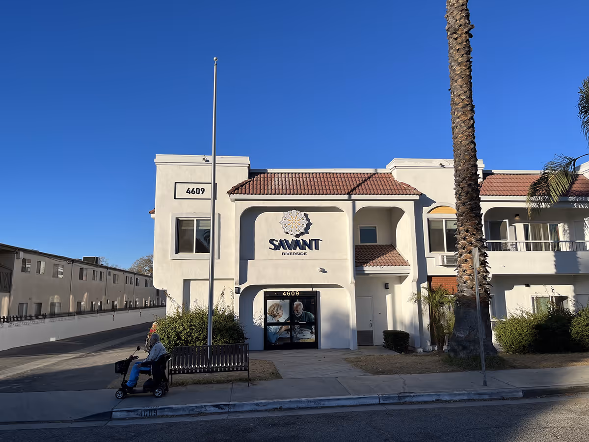Exterior front view of Savant of Riverside senior living facility building with white walls and red tile roof under a clear blue sky. A person on a mobility scooter is on the sidewalk near a bench and a tall palm tree is visible on the right side.
