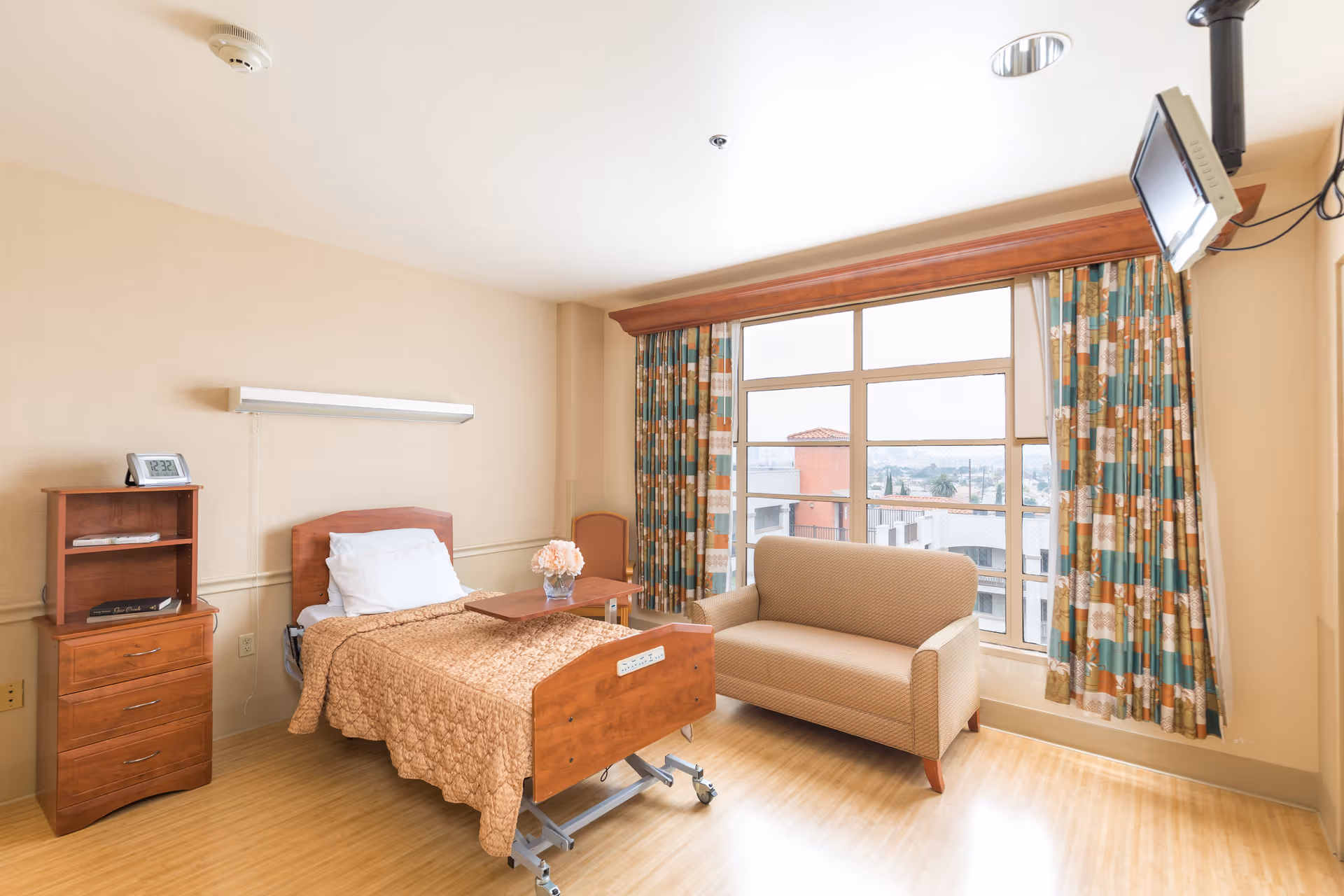 A bright and clean patient room with a hospital bed covered in a brown quilt, a small wooden nightstand with drawers and a clock, a beige loveseat, a wooden chair, and large windows with patterned curtains letting in natural light. A TV is mounted on the wall near the ceiling.