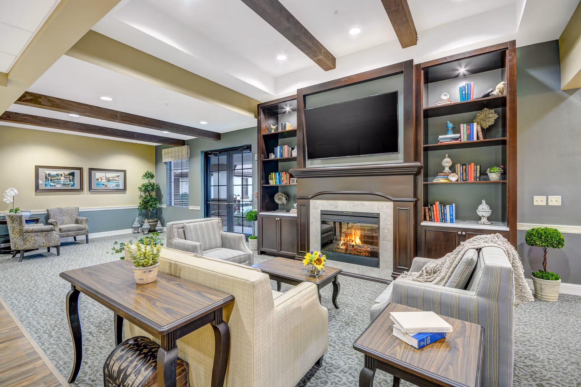 A cozy living room area in Tuscan Gardens Of Venetia Bay featuring a fireplace with a mounted TV above it, flanked by built-in dark wood bookshelves filled with books and decorative items. The room has comfortable seating including a beige sofa, striped armchairs, and wooden tables with plants and books. The ceiling has exposed wooden beams and recessed lighting, and there is a glass door leading outside.