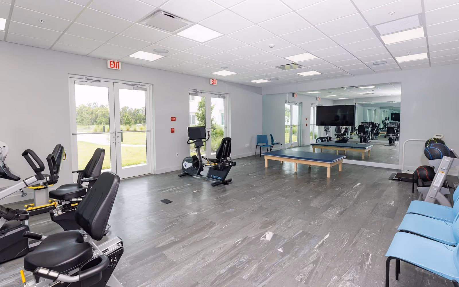 A spacious fitness room in a senior living facility with exercise bikes, a large mirror on one wall, a flat-screen TV, exercise balls, and blue chairs along the right side. The room has large windows and glass doors letting in natural light and showing a green outdoor area.