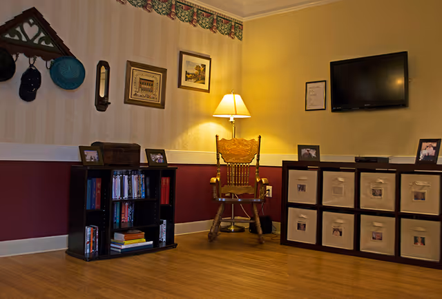 A cozy living room area with wooden flooring, a wooden rocking chair illuminated by a floor lamp, a small bookshelf filled with books and DVDs, and a storage unit with fabric bins. The walls are decorated with framed pictures, a mounted TV, and a decorative hat rack with hats hanging on it.