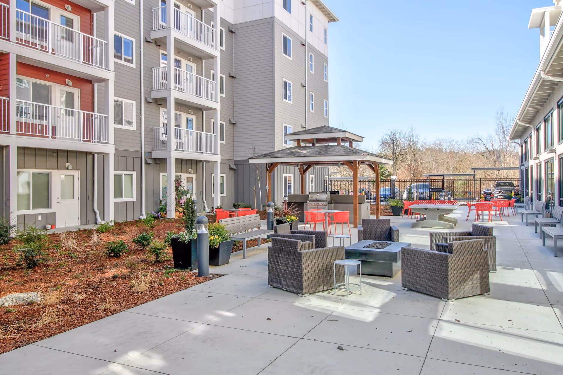 Outdoor patio area at the Reserve at Auburn featuring modern wicker chairs arranged around a fire pit, red chairs and tables, a gazebo with a grill, benches, and landscaped garden beds next to a multi-story residential building under a clear blue sky.